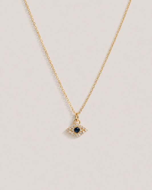A delicate gold chain necklace featuring an evil eye pendant with a central dark blue sapphire surrounded by small diamonds, displayed on a light beige background.