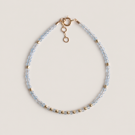 A delicate beaded bracelet with aquamarine faceted beads and small gold spacer beads, featuring a gold spring ring clasp and extension chain, displayed on a neutral beige background.