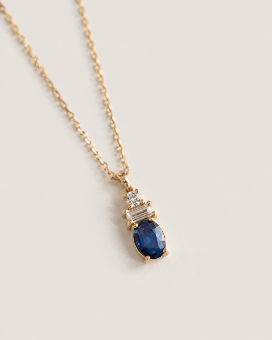 A gold chain necklace featuring a pendant with a large oval deep blue gemstone and smaller clear rectangular stones above it, set against a light beige background.